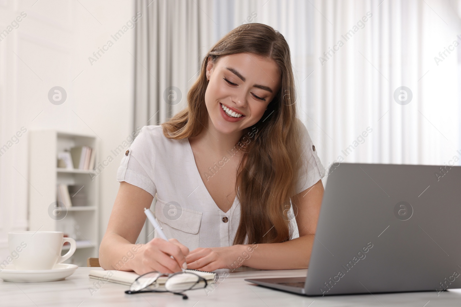 Photo of Happy woman writing something in notebook near laptop at white table in room