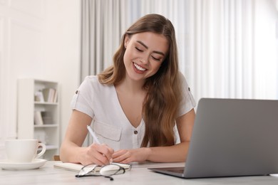 Happy woman writing something in notebook near laptop at white table in room