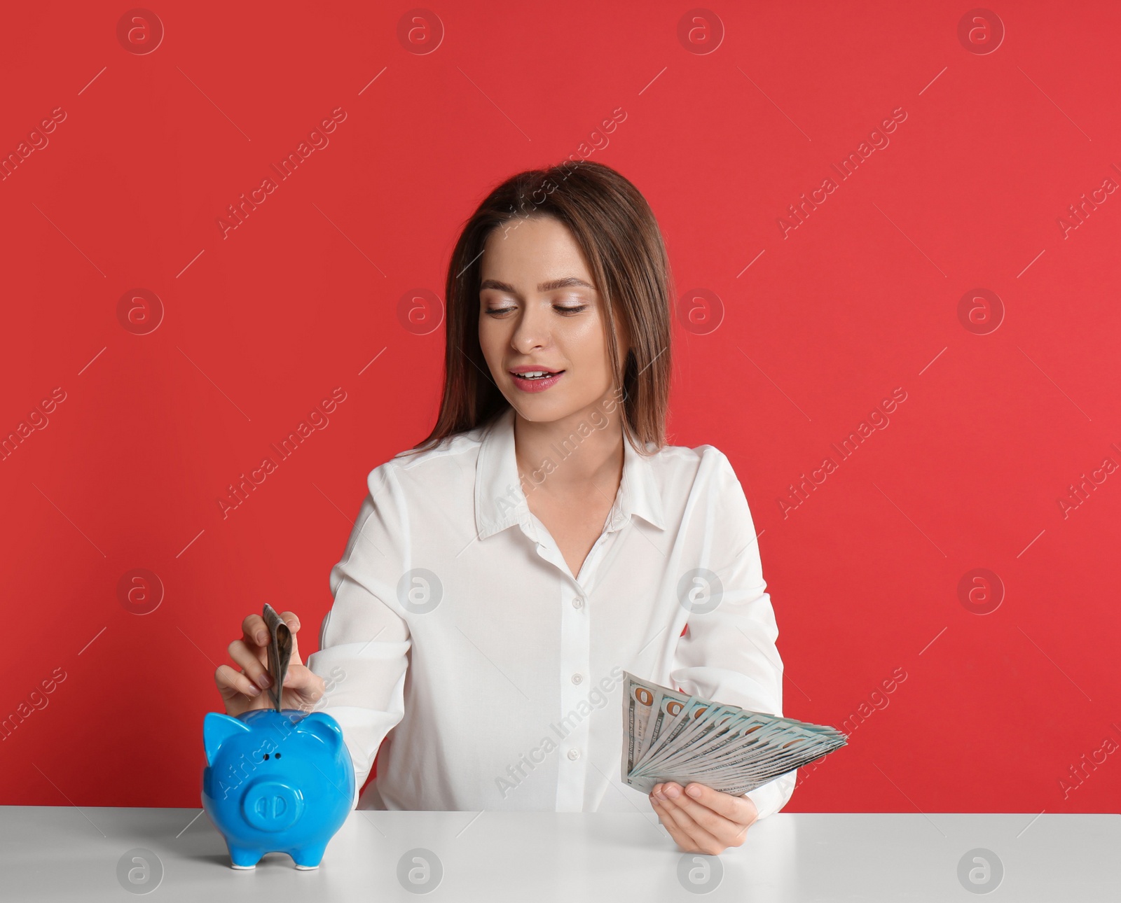Photo of Young woman putting money into piggy bank at table on crimson background