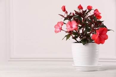 Beautiful impatiens flower in pot on white wooden table near light wall, space for text