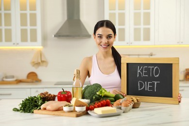 Happy woman holding chalkboard with words Keto Diet near different products in kitchen