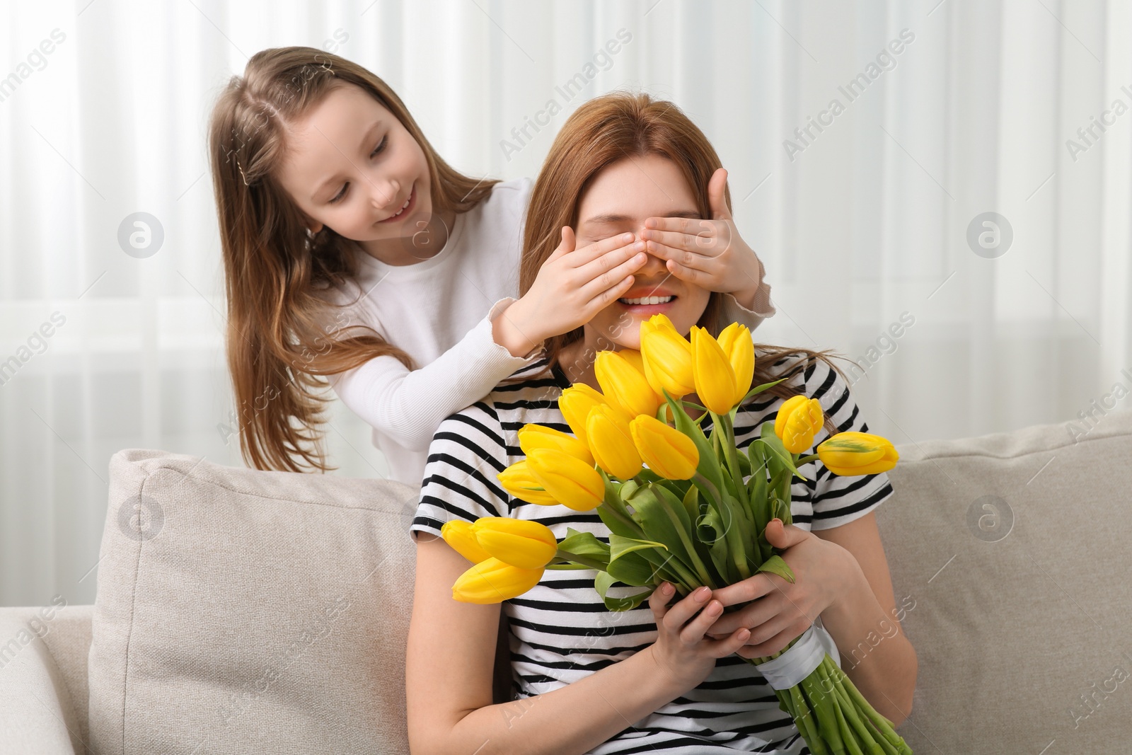 Photo of Daughter covering mother's eyes with her palms and congratulating with bouquet of yellow tulips at home