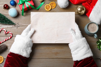 Photo of Santa Claus holding empty paper at wooden table, top view. Space for text