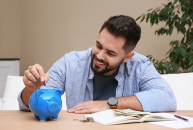 Photo of Young man putting coin into piggy bank at table indoors. Money savings