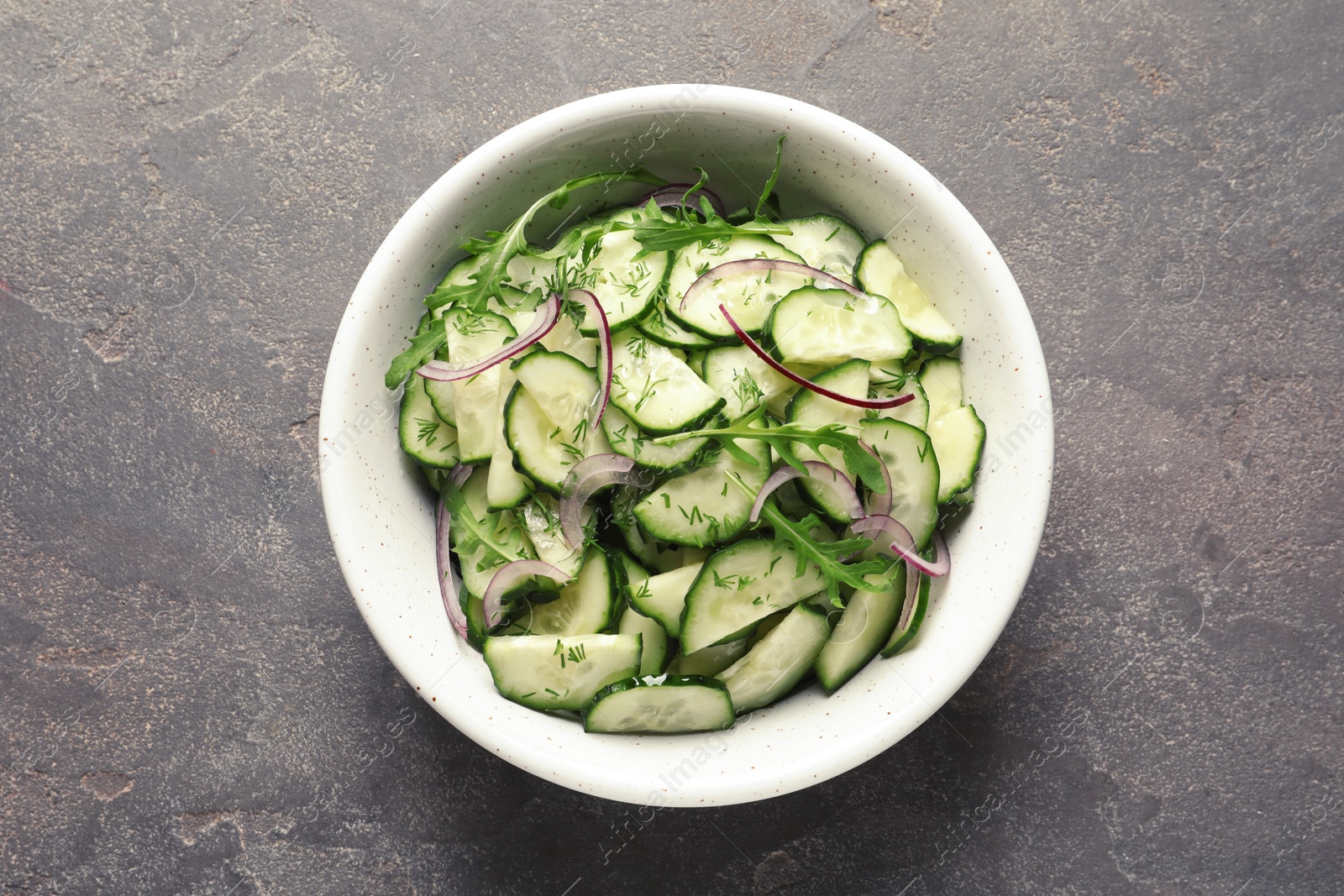 Photo of Delicious cucumber salad with onion and arugula in bowl on grey background, top view