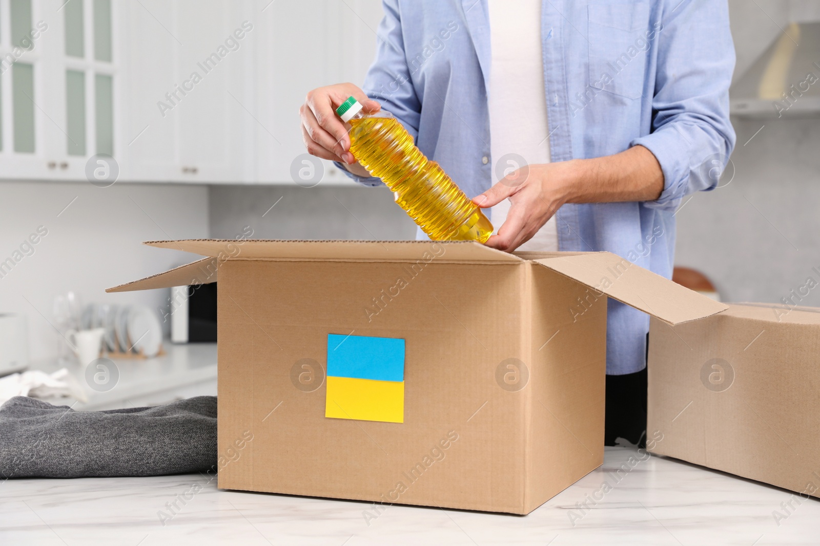 Photo of Humanitarian aid for Ukraine. Man putting bottle of oil in donation box at white table indoors, closeup