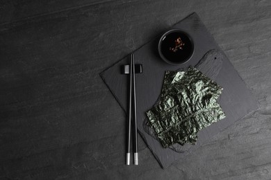 Photo of Dry nori sheets, soy sauce and chopsticks on black table, flat lay. Space for text