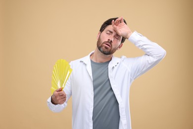 Unhappy man with hand fan suffering from heat on beige background