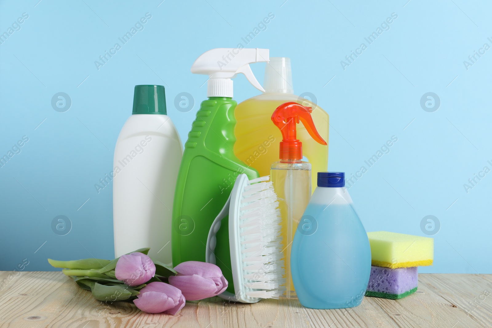 Photo of Spring cleaning. Different detergents, tools and beautiful flowers on wooden table against light blue background