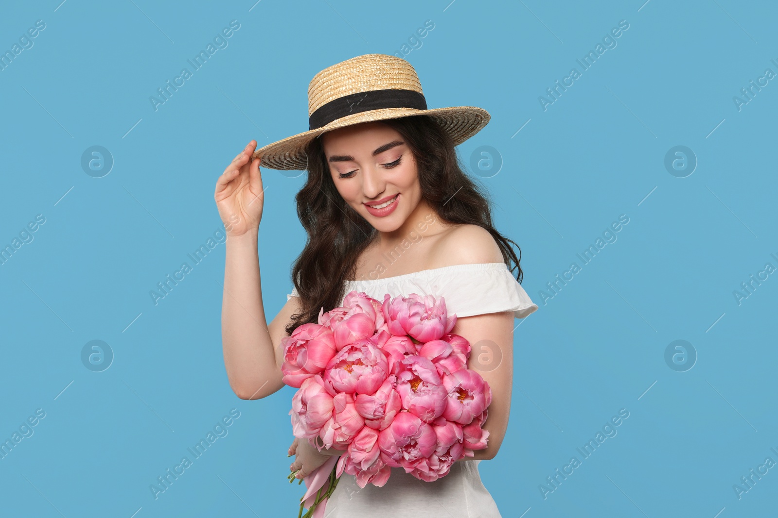 Photo of Beautiful young woman in straw hat with bouquet of pink peonies against light blue background