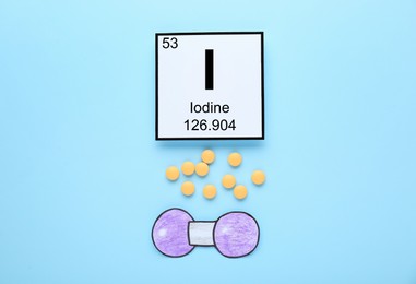 Photo of Card with iodine element, atom made of paper and pills on light blue background, flat lay