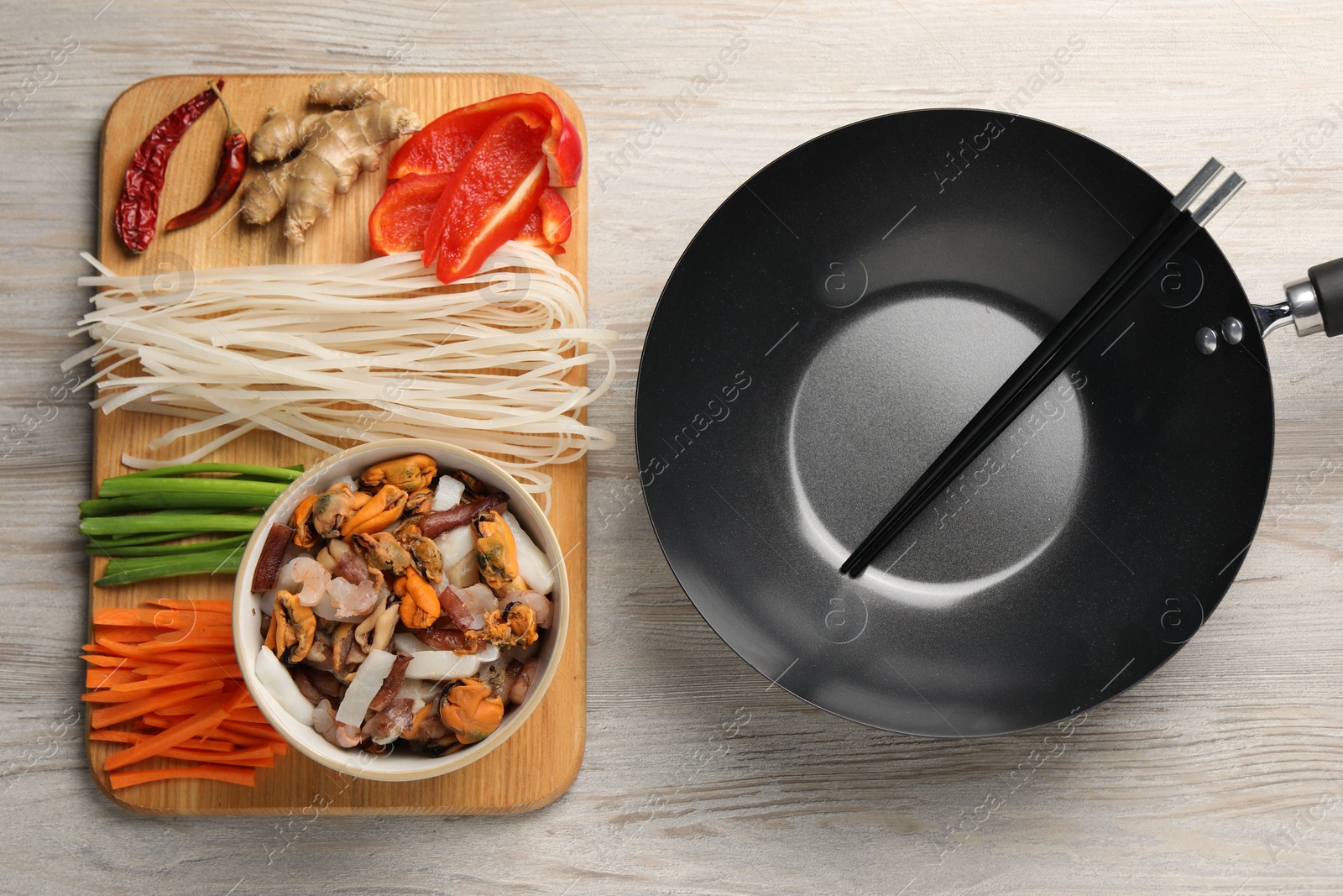 Photo of Black wok, chopsticks and board with products on light wooden table, top view