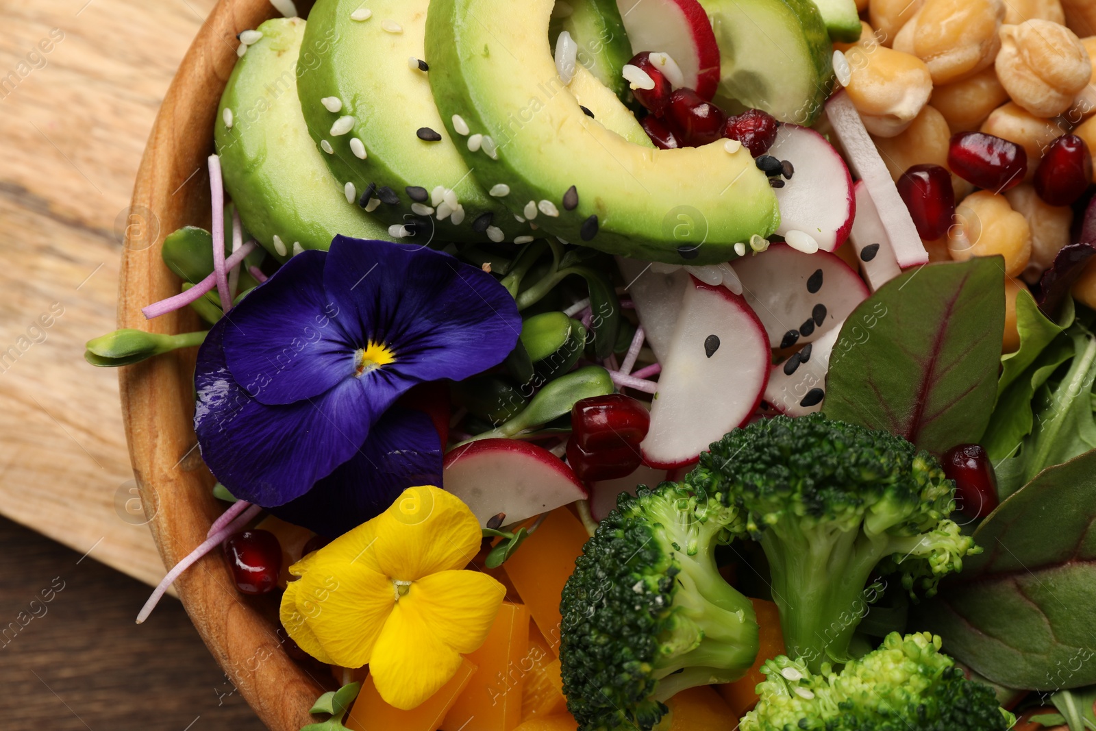 Photo of Delicious vegan bowl with broccoli, avocados and violet flowers on wooden table, top view