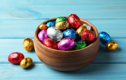 Photo of Bowl with chocolate eggs wrapped in colorful foil on light blue wooden table
