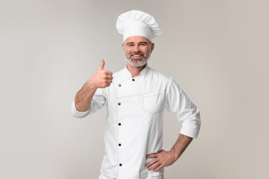 Happy chef in uniform showing thumbs up on grey background