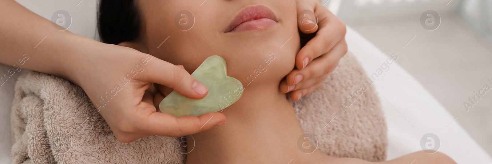 Image of Young woman receiving facial massage with gua sha tool in beauty salon. Banner design