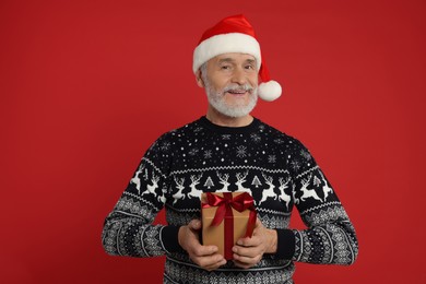 Senior man in Christmas sweater and Santa hat holding gift on red background