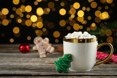Delicious hot chocolate with marshmallows and Christmas tree gingerbread cookie on wooden table against blurred lights, space for text