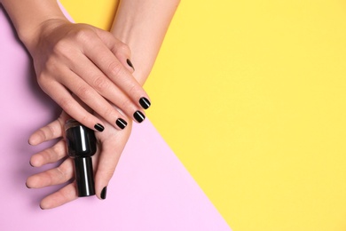 Woman with black manicure holding nail polish bottle on color background, top view. Space for text