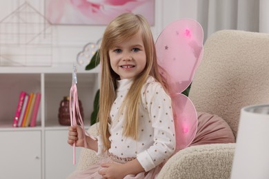 Photo of Cute little girl in fairy costume with pink wings and magic wand in armchair at home