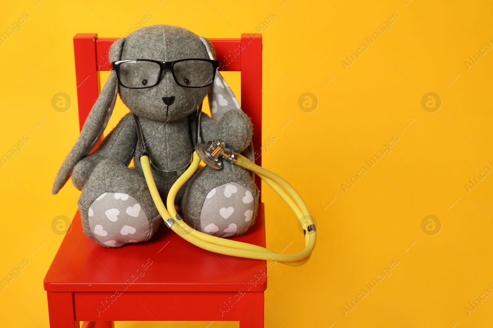 Photo of Toy bunny with stethoscope and glasses on chair against yellow background, space for text. Pediatrician practice