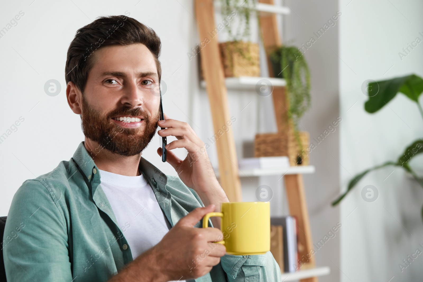 Photo of Handsome man with cup of drink talking on phone at home, space for text