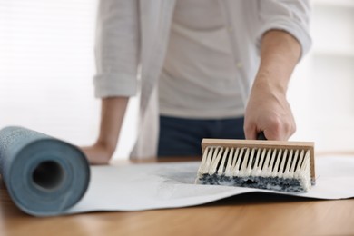 Photo of Man applying glue onto wallpaper sheet at table indoors, closeup