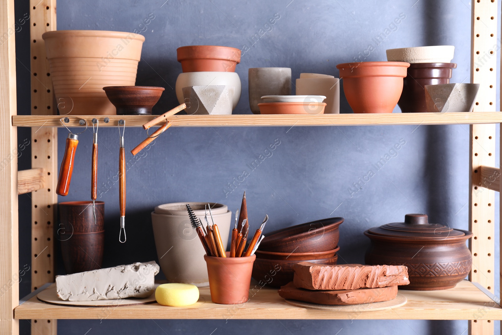 Photo of Set of different crafting tools and clay dishes on wooden rack in workshop