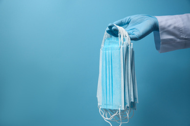 Photo of Doctor in latex gloves holding disposable face masks on light blue background, closeup with space for text. Protective measures during coronavirus quarantine