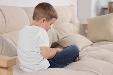 Photo of Boy with poor posture sitting on beige sofa in room. Symptom of scoliosis
