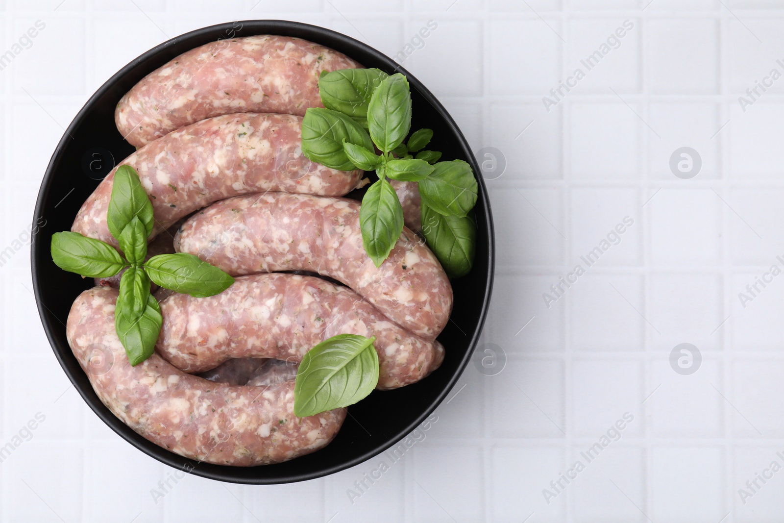 Photo of Raw homemade sausages and basil leaves on white tiled table, top view. Space for text