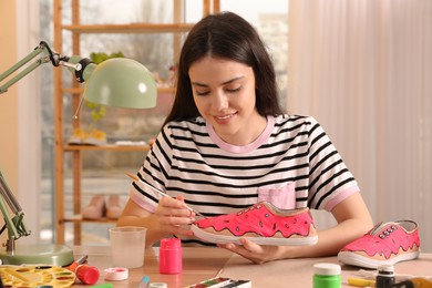 Photo of Woman painting on sneaker at wooden table indoors. Customized shoes