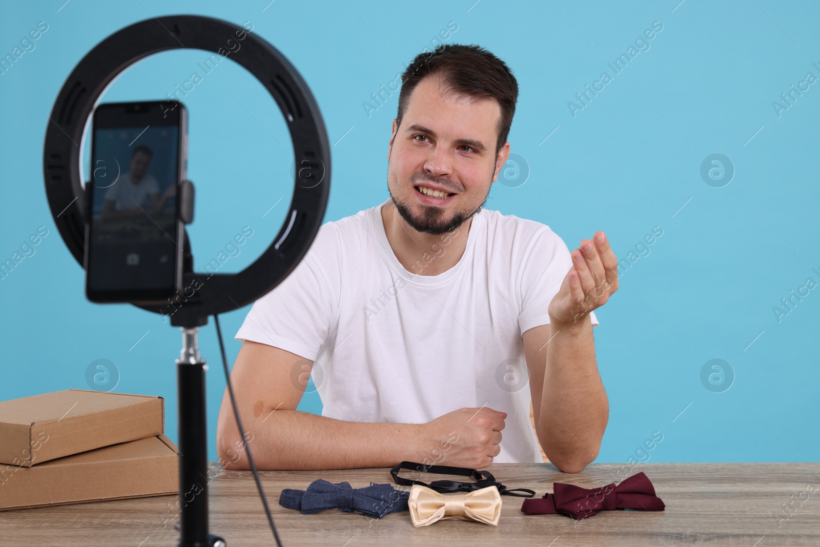 Photo of Smiling fashion blogger showing bow ties while recording video at table against light blue background