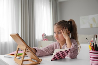 Photo of Bored little girl doing homework with tablet at table indoors