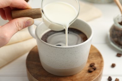 Woman pouring milk into cup with coffee at white table, closeup