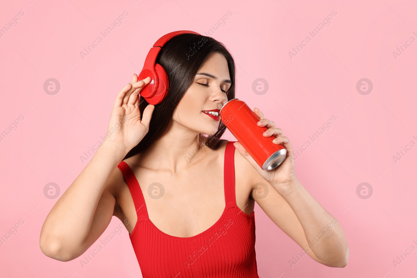 Photo of Beautiful young woman with headphones drinking from tin can on pink background