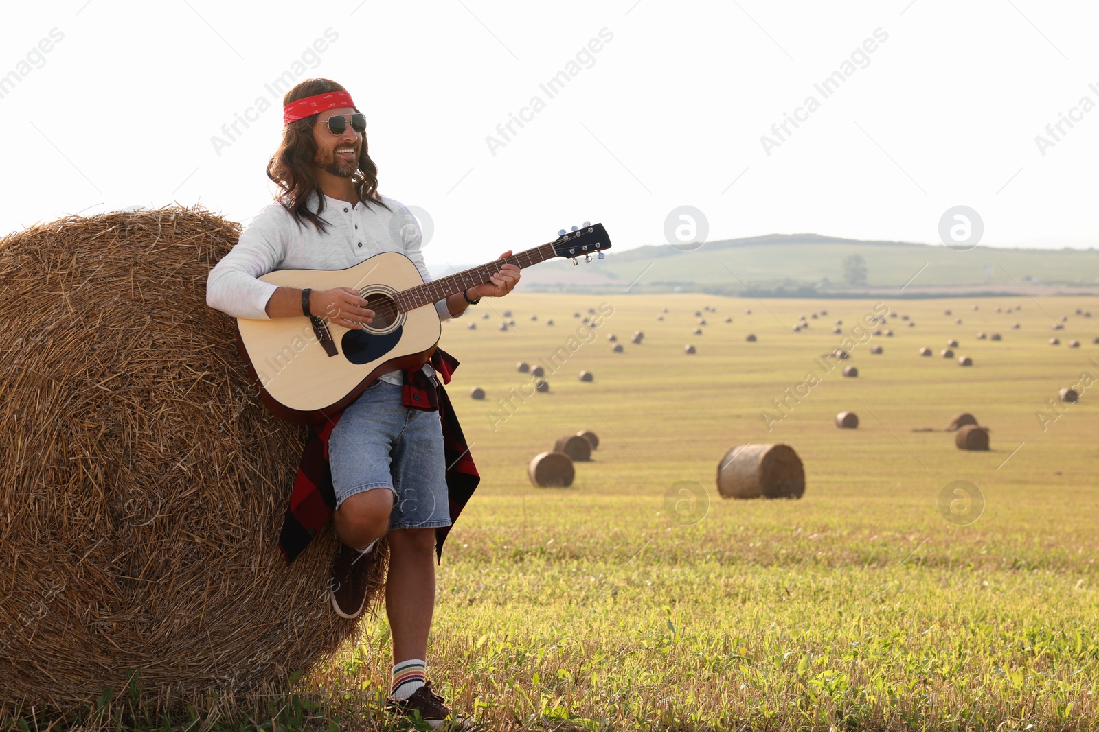 Photo of Hippie man playing guitar near hay bale in field, space for text