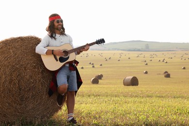 Hippie man playing guitar near hay bale in field, space for text