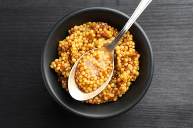 Whole grain mustard in bowl and spoon on black wooden table, top view