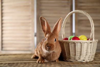 Photo of Cute bunny and basket with Easter eggs on wooden table against blurred background