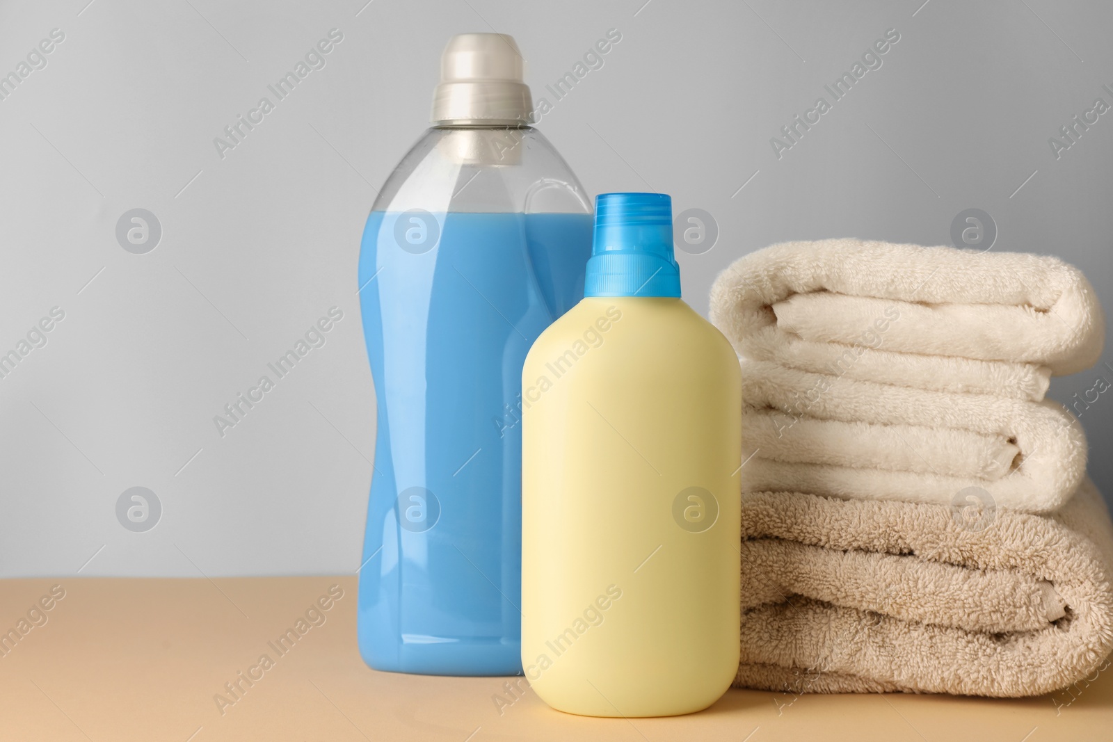 Photo of Bottles of fabric softener and stacked clean towels on pale yellow table against light grey background, space for text