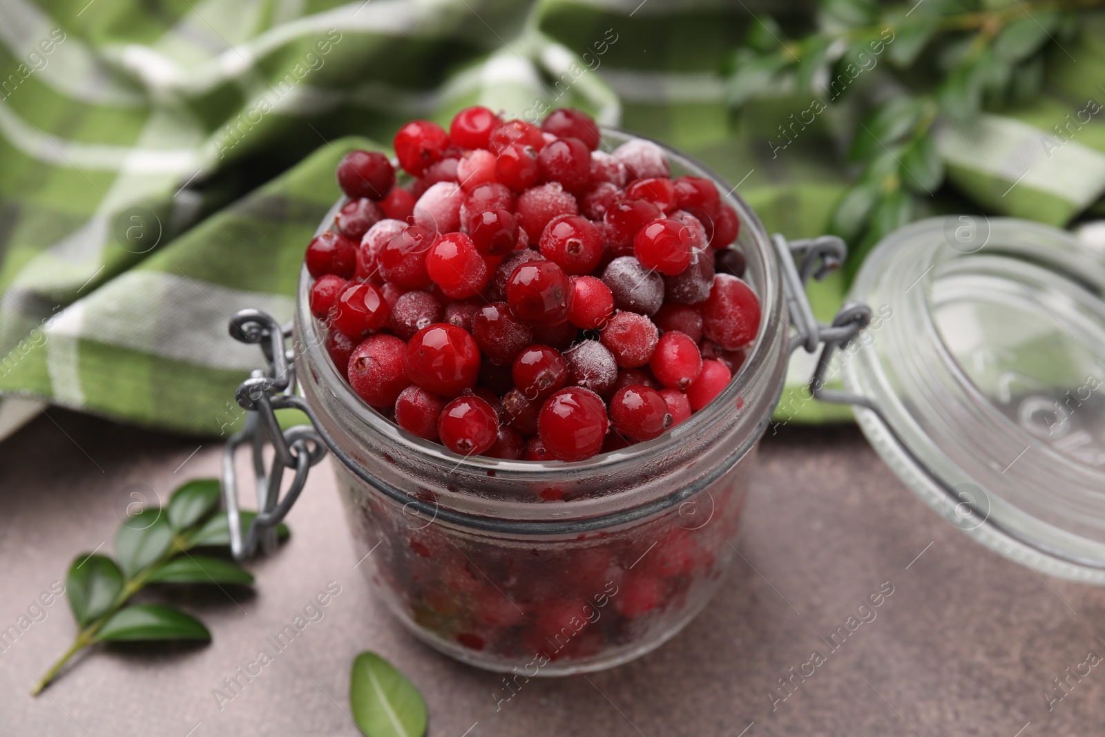 Photo of Frozen red cranberries in glass jar and green leaves on brown textured table, closeup