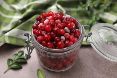 Frozen red cranberries in glass jar and green leaves on brown textured table, closeup