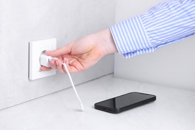 Woman plugging smartphone into power socket at white table indoors, closeup