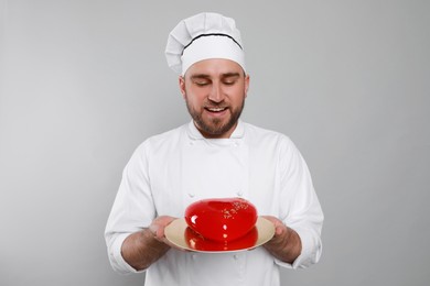 Happy professional confectioner in uniform holding delicious cake on light grey background