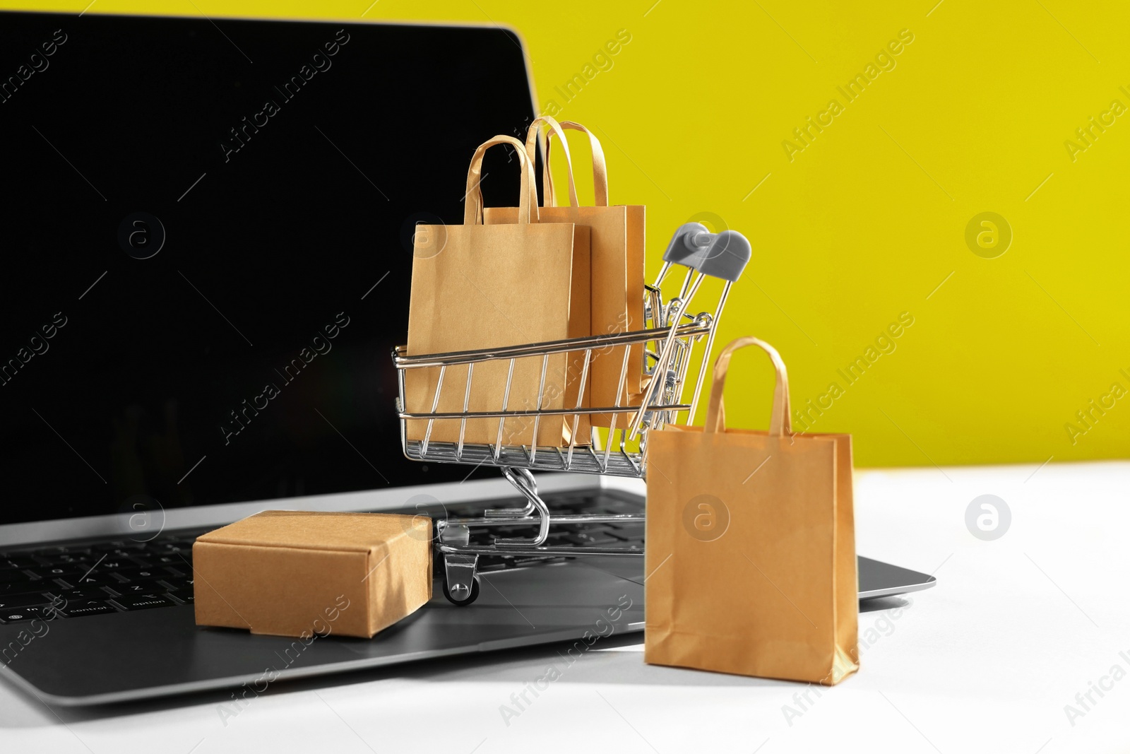 Photo of Online store. Laptop, mini shopping cart and purchases on beige table