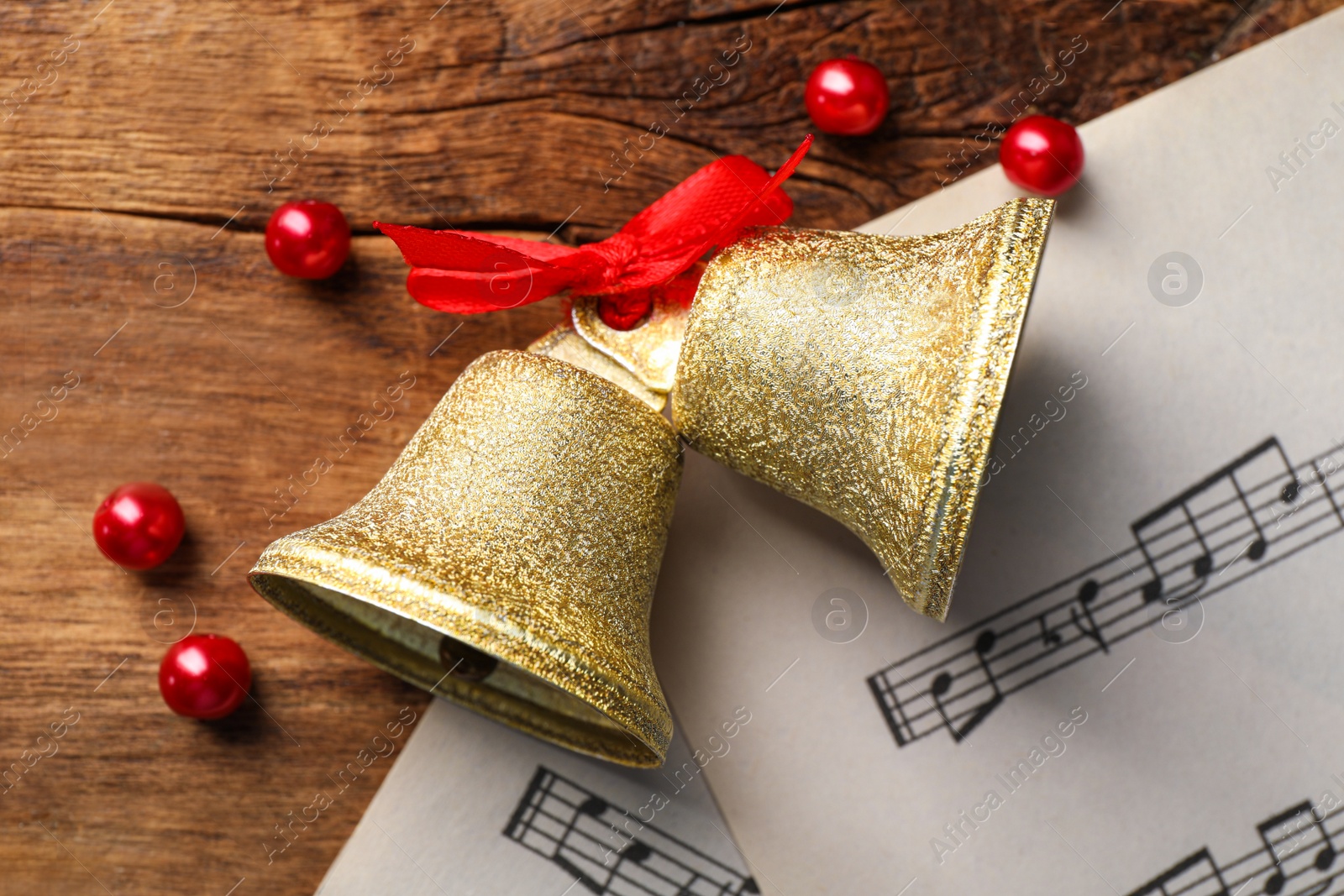 Photo of Golden shiny bells with red bow and music sheets on wooden table, flat lay. Christmas decoration