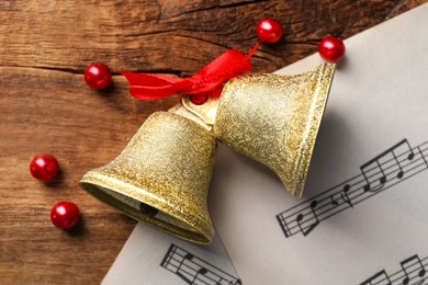 Photo of Golden shiny bells with red bow and music sheets on wooden table, flat lay. Christmas decoration
