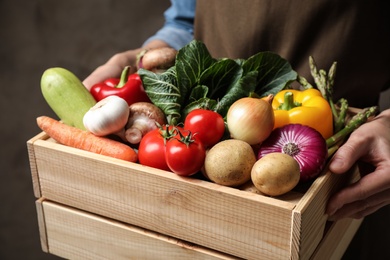 Photo of Man holding wooden crate full of fresh vegetables on brown background, closeup