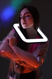 Fashionable portrait of beautiful woman wearing transparent coat with square lamp on dark background in neon lights